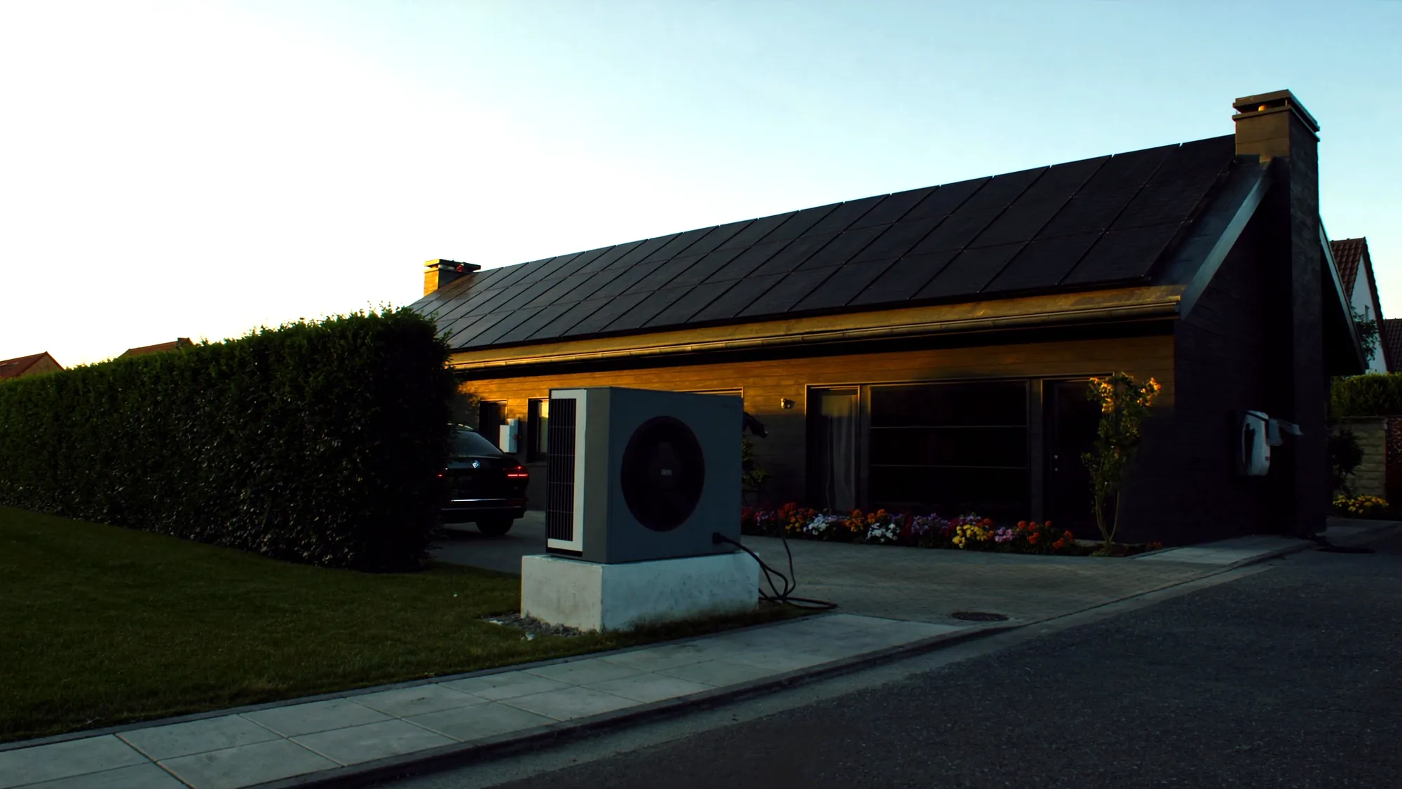 Einfamilienhaus mit vollflächigem Solardach, Wärmepumpe neben dem Haus und Auto am Carport im Abendlicht